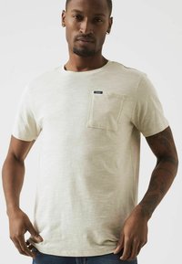 Camiseta de manga corta beige claro en un tejido suave y texturizado, con un bolsillo en el pecho que tiene una pequeña etiqueta en azul marino, cuello redondo y un corte relajado.