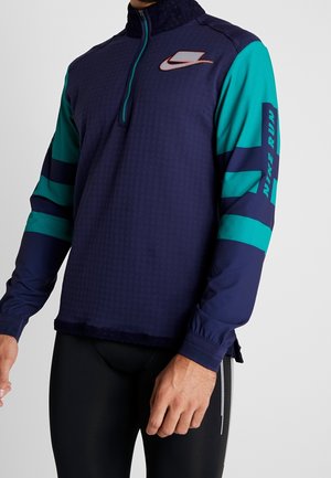 Mand iført en navy- og tealfarvet langærmet Nike løbetrøje med kvart lynlås og logo på bryst og ærme, kombineret med sorte atletikleggings.