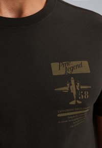 Camiseta de algodón negra con cuello redondo, que presenta un diseño impreso en verde oliva, incluyendo el texto "Pme Legend" y un gráfico de un avión.