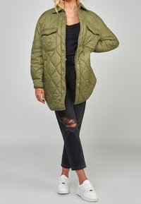 Chaqueta acolchada de color verde oliva, de corte oversize, con cierre de botones, dos bolsillos frontales y jeans negros desgastados, combinados con zapatillas blancas.