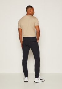 Beige rövidujjú póló sötétkék slim-fit nadrággal, amelyen két hátsó zseb és gombok találhatók, fehér sportcipővel viselve.