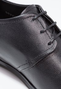 Chaussure en cuir noir avec une surface texturée, présentant une finition lisse, un éclat subtil et des lacets assortis. Vue en gros plan de la partie supérieure.