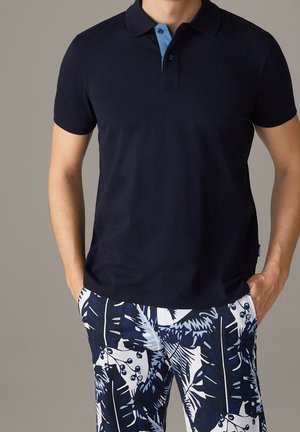 Mand iført en marineblå polo med en lyseblå indvendig krave og blå-hvide tropicalmønstrede shorts, hænderne i lommerne.