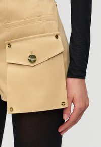 Μπεζ σορτς με μεγάλη τσεπάκη με καπάκι και χρυσά κουμπιά, φορεμένα πάνω από μαύρο καλσόν, με ορατό χέρι και μαύρο μακρυμάνικο μπλουζάκι.