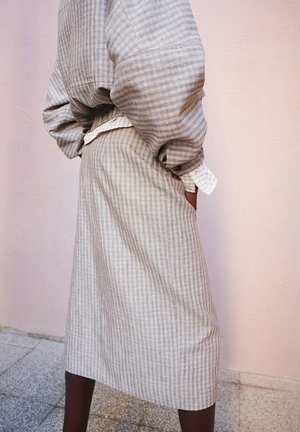 Osoba odjevena u bež kariranu dugu suknju i odgovarajuću široku košulju s rukom u džepu, stoji naslonjena na blijedoružičasti zid.