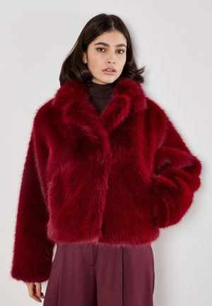 Rode nep bontjas met een brede kraag, dikke textuur en een oversized silhouet, gecombineerd met een donkere top en losse broek.