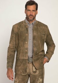 Bruine suede jas met een staande kraag, metalen knopen en een borstzak, gedragen over een zwart-wit geruite shirt.