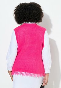 Fuchsia gebreide mouwloze vest met een gestructureerde afwerking, met een franjerand, gedragen over een witte blouse.