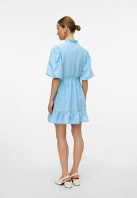 Svetlo modra obleka z naborkom na robu, napihnjenimi rokavi in ustreznim pasom, izdelana iz lahkotnega materiala. Nیزodeta z belimi čevlji na blok petah.