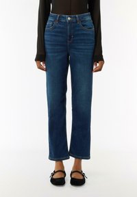Dunkelblaue Jeans mit hohem Bund, geradem Schnitt, fünf Taschen und kontrastierenden orangen Nähten. Kombiniert mit schwarzen flachen Schuhen.