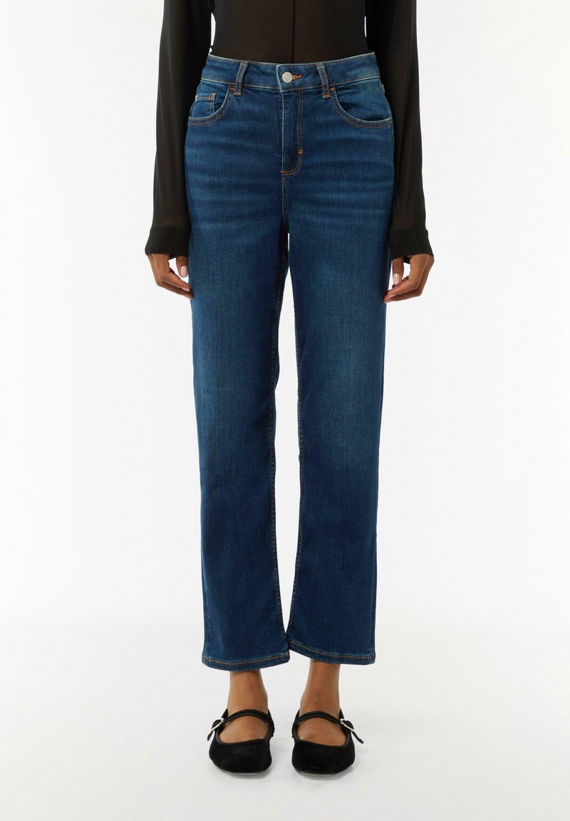 Dunkelblaue Jeans mit hohem Bund, geradem Schnitt, fünf Taschen und kontrastierenden orangen Nähten. Kombiniert mit schwarzen flachen Schuhen.