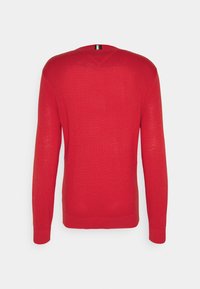 Pull en maille rouge avec col en V, manches longues, poignets et ourlet côtelés, et une étiquette rayée contrastante à l'arrière du col.