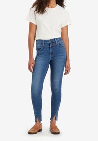 Jeans ajustados de mezclilla azul medio, con cintura alta, dos bolsillos frontales y aberturas laterales en el dobladillo. Combinados con una camiseta blanca y mocasines marrones.