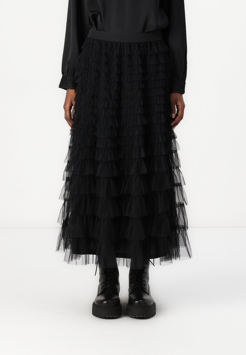 Personne portant une jupe noire à volants superposés associée à des bottines noires à lacets et plateforme, debout devant un fond blanc uni.