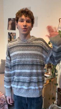 Młoda osoba o kręconych włosach, ubrana w szary sweter w paski, macha prawą ręką w przytulnym pokoju z roślinami i dekoracjami na ścianie.