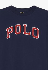 Marinblå bomullströja med "POLO" i stora röda strukturerade bokstäver med vita accenter på bröstet. Rund halsringning.