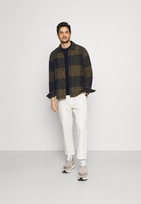 Cămașă flanelată în carouri, în nuanțe de măslin și albastru închis, purtată cu un top albastru, pantaloni albi și adidași gri cu accente subtile, stând pe un fundal simplu.