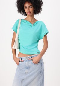 Teal bomba iz bombažnega materiala z zvitim detajlom na vratu in kratkimi rokavi, v kombinaciji z svetlo modrim jeans krilom in majhno bež torbico.