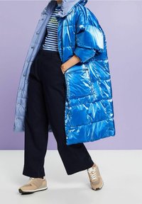 Skinande blå puffjacka med huva och stor ficka, kombinerad med en randig blå och vit topp, svarta vida byxor och beige sneakers.