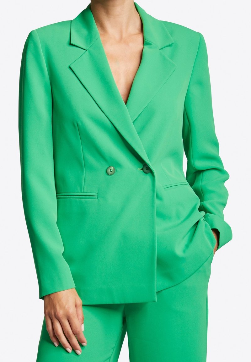 Green tailored blazer with a double-breasted design, notched lapels, and pockets; made from smooth fabric with a vibrant solid color.