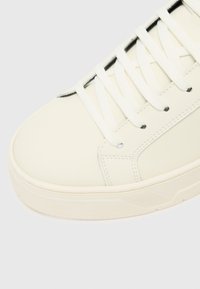 Weiße Leder-Sneaker mit flachen Schnürsenkeln, abgerundetem Zehenbereich, glatter Textur und minimalen Akzenten. Sichtbare Steppnähte an den Seiten und auf der Oberfläche.