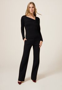 Zwarte long sleeve top met een asymmetrische halslijn en knoopdetail, gecombineerd met strakke zwarte broek en bordeauxrode puntige hakken.