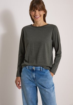 Femme souriante portant un t-shirt rayé à manches longues gris foncé et un jean bleu clair, une main dans la poche, debout devant un fond uni.