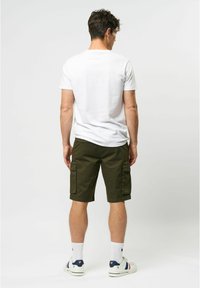 Tricou alb cu manșete scurte, pantaloni scurți cargo de culoare verde măslin cu două buzunare laterale, până la genunchi, asortați cu șosete albe și adidași.