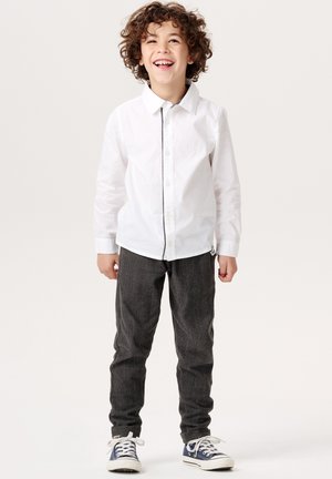 Camicia bianca a maniche lunghe, pantaloni grigi a fantasia e sneakers blu. Un bambino sorride, con capelli ricci e mani rilassate ai lati.