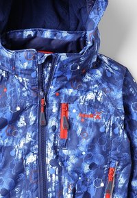 Veste de neige bleue avec un design à motifs et des accents hexagonaux. Comprend des tirettes de fermeture éclair rouges et plusieurs poches. Tissu texturé sur l'ensemble.