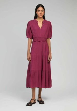 Robe longue de couleur prune avec un décolleté en V, des manches bouffantes et une jupe à volants. Présente une taille froncée et des détails subtils le long de l'encolure.