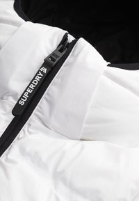 Chaqueta blanca acolchada con un zipper negro y el logotipo "SUPERDRY". Presenta un diseño acolchado y un cuello alto, destacando un diseño moderno y aislante.