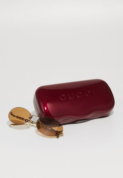 Punainen kiiltävä aurinkolasikotelo, jossa on kohokuvioitu "GUCCI"-logo. Mukana ovaalinmuotoiset aurinkolasit ruskeilla linsseillä ja ketjuilla koristeltu kehys.