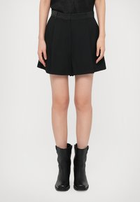 Benen van een persoon in zwarte geplooide shorts, zwarte leren laarzen tot halverwege de kuit, staande tegen een witte achtergrond.