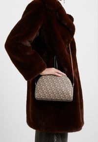 Beigen kuvioitu käsilaukku, jossa on kaareva muoto, mustat reunat ja ruskea hihna, pidetään ruskean pörröisen takin päällä. Takissa on suuri kaulus.