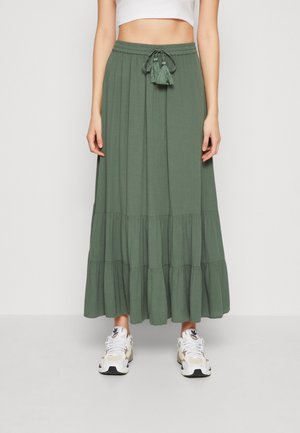 Jupe longue olive en tissu léger, dotée d'une taille avec cordon de serrage et des pompons, ainsi qu'un design en volants superposés. Portée avec des baskets blanches.