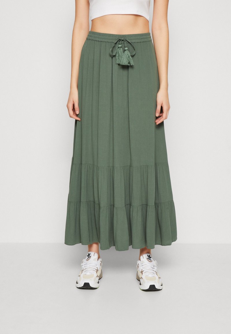 Jupe longue olive en tissu léger, dotée d'une taille avec cordon de serrage et des pompons, ainsi qu'un design en volants superposés. Portée avec des baskets blanches.