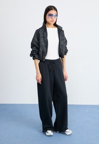 Un modelo se presenta con confianza, vistiendo una chaqueta bomber negra con diseño, una camiseta blanca y pantalones negros holgados, con gafas de sol azules, frente a un fondo claro.