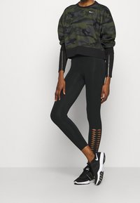 Sweat-shirt court à motif camouflage vert foncé, leggings noirs avec découpes, et chaussures de sport noires avec logo blanc.