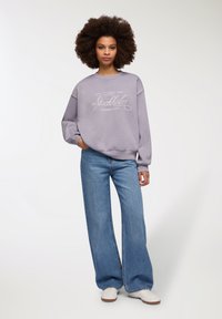 Sweatshirt di un leggero viola con grafiche bianche che recitano "Stockholm Leisure Club" abbinato a jeans in denim a vita alta e gamba larga e sneaker bianche.