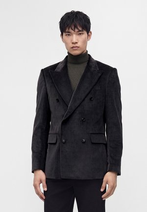Homme portant un blazer en velours côtelé gris foncé à double boutonnage sur un pull à col roulé vert olive, debout devant un fond clair uni.