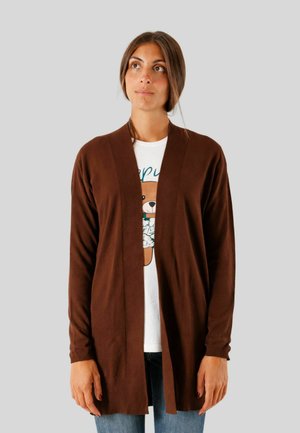 Donna che indossa un cardigan marrone aperto davanti sopra una t-shirt bianca con una grafica di un orsetto, abbinata a jeans blu, in piedi contro uno sfondo semplice.