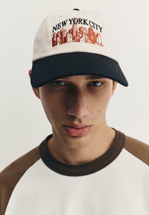Baseballkappe aus cremefarbenem Stoff mit schwarzem Schirm, versehen mit rotem "NEW YORK CITY"-Text und Skyline-Grafik. Aus Baumwollmaterial mit strukturierter Form.