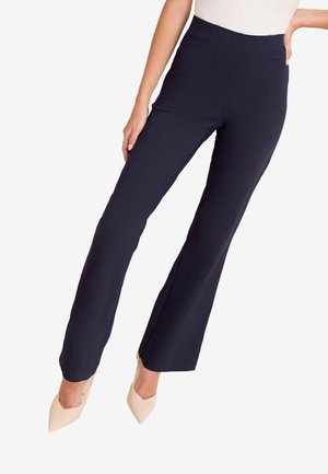 Kvinde iført tætsiddende marineblå bootcut-bukser og beige spidse hæle, stående med den ene hånd på hoften.