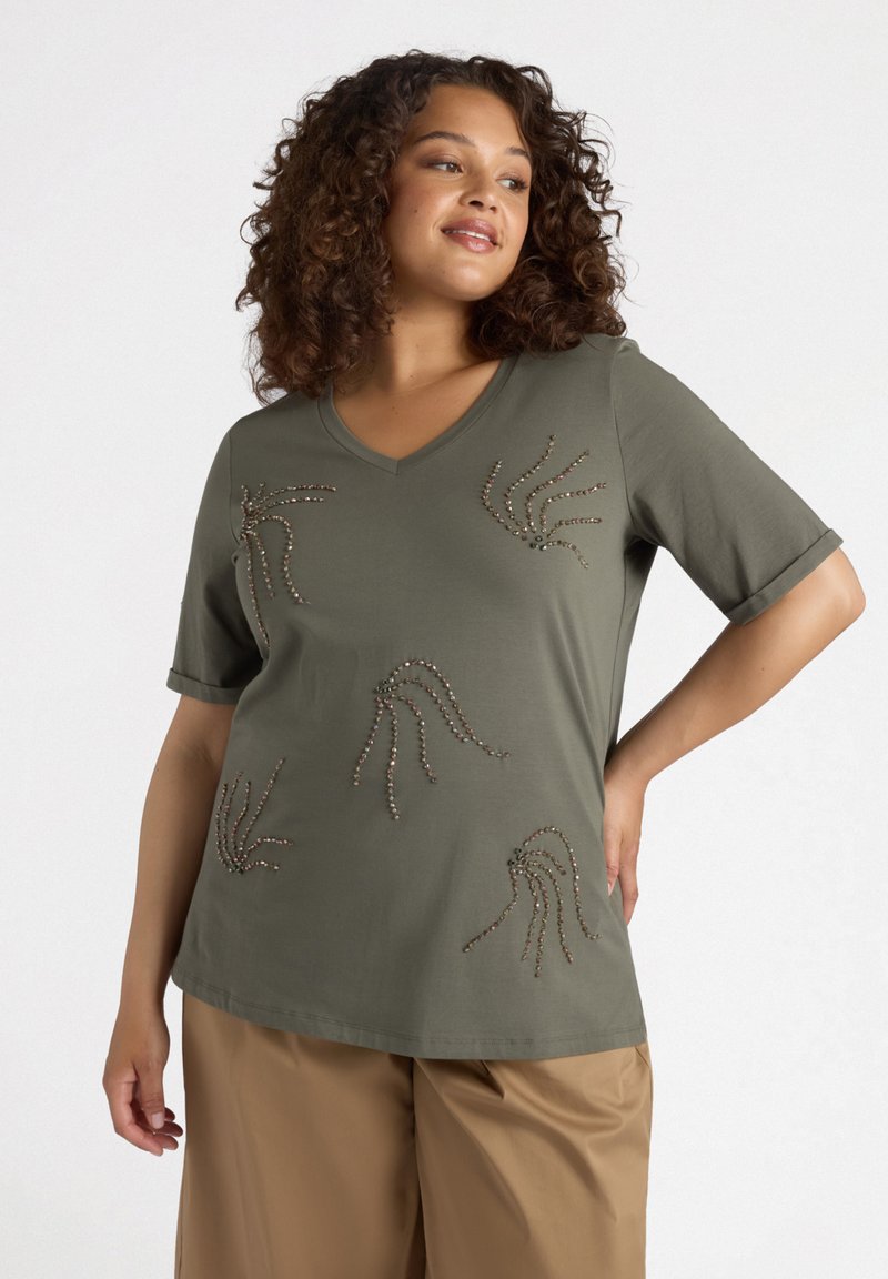 Camiseta de color verde olivo con escote en V y mangas cortas, con diseños de ondas de cuentas en varios colores en la parte delantera, combinada con pantalones beiges.