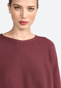 Haut en tricot bordeaux avec un col rond et un tissu texturé. Le design présente des détails de couture subtils et une silhouette ample.