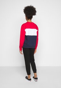 Persoon met krullend haar, gekleed in een rood-wit-navy color-block sweatshirt, zwarte broek en zwarte sneakers, staande met de rug naar voren.