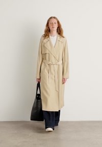 Beige Trenchcoat mit breiten Revers, betontem Taillengürtel und seitlichen Taschen, kombiniert mit einer dunklen Jeanshose und einer schwarzen Tragetasche.