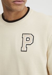 Sudadera crema con un cuello redondo de canalé marrón. Presenta una letra "P" bordada y texturizada en beige y negro en la parte frontal.