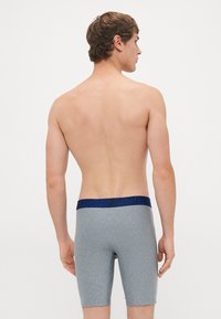 Uomo visto di spalle che indossa boxer grigi con un elastico blu scuro, in piedi contro uno sfondo bianco semplice.